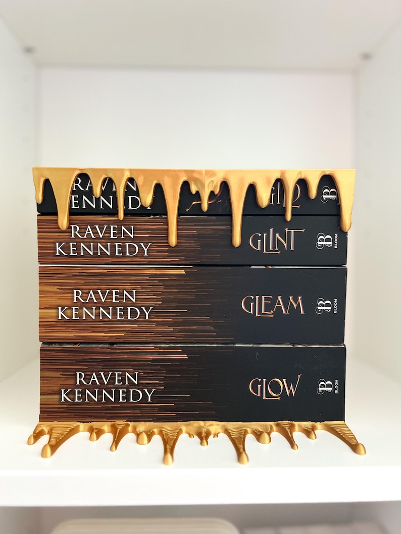 May include: A stack of three books with black covers and gold lettering, titled "Glint", "Gleam", and "Glow" by Raven Kennedy. The books are held by a gold-colored decorative shelf with a dripping paint design.