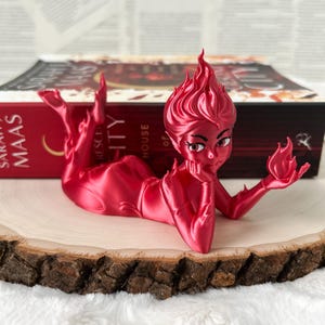 May include: A red figurine of a character with flame-like hair and a thoughtful expression, holding a small flame. The figure is lying on a wooden slice, with a book in the background. The figurine is a unique decorative item.