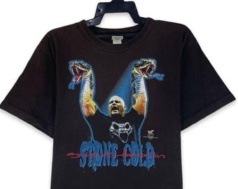 May include: A black short-sleeve t-shirt featuring a graphic of a wrestler with arms raised, flanked by two snakes. The text "STONE COLD" appears in blue lettering below the figure, with a small WWE logo visible on the right side of the graphic.