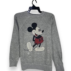 Puede incluir: Sudadera gris con un gráfico de Mickey Mouse. El personaje de dibujos animados está representado en negro, rojo y blanco. La sudadera tiene mangas largas y cuello redondo, colgada de una percha negra. Diseño clásico de Disney.