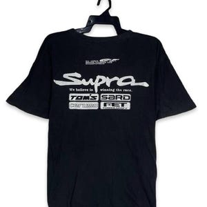 May include: A black short-sleeved t-shirt featuring white graphic text that reads Supra, We believe in winning the race, TOM'S, SARD, cerumo, and FET. The shirt includes a small All Japan Grand Touring Car Championship logo at the top center.