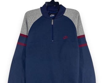 May include: A navy blue Nike sweatshirt with a gray yoke and sleeves. The sleeves have a maroon stripe. The sweatshirt has a quarter-zip closure and a small Nike logo on the chest. The sweatshirt is hanging on a black hanger.