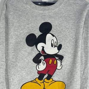 Puede incluir: Una sudadera gris con un gran gr&aacute;fico de Mickey Mouse. Mickey se muestra con su atuendo cl&aacute;sico: pantalones cortos rojos, zapatos amarillos, guantes blancos y cuerpo y orejas negros. La sudadera tiene cuello redondo y mangas largas.
