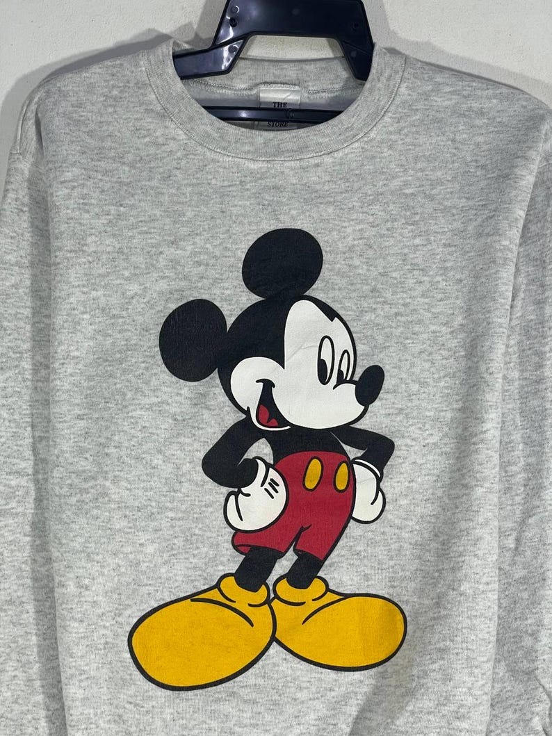 Puede incluir: Una sudadera gris con un gran gr&aacute;fico de Mickey Mouse. Mickey se muestra con su atuendo cl&aacute;sico: pantalones cortos rojos, zapatos amarillos, guantes blancos y cuerpo y orejas negros. La sudadera tiene cuello redondo y mangas largas.