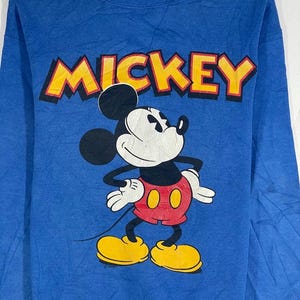 以下が含まれることがあります： ミッキーマウスが描かれた青いスウェットシャツ。「MICKEY」の文字が黒い縁取りの黄色でプリントされています。ミッキーマウスは、赤いパンツと黄色の靴を履いたクラシックなポーズで描かれています。