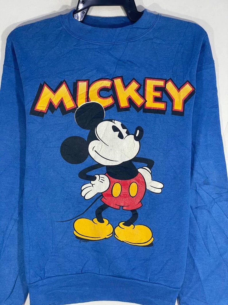以下が含まれることがあります： ミッキーマウスが描かれた青いスウェットシャツ。「MICKEY」の文字が黒い縁取りの黄色でプリントされています。ミッキーマウスは、赤いパンツと黄色の靴を履いたクラシックなポーズで描かれています。