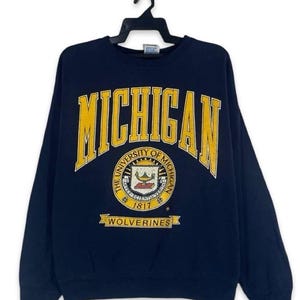 Può includere: Una felpa blu navy a girocollo con la scritta MICHIGAN in grandi lettere gialle sul petto. Sotto il testo si trova il sigillo dell'Università del Michigan con la data 1817 e un banner giallo contenente la parola WOLVERINES.
