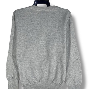 Puede incluir: Una sudadera gris jaspeado con cuello redondo y mangas largas. Los pu&ntilde;os y el dobladillo son acanalados. La sudadera est&aacute; colgada de una percha negra sobre un fondo blanco. Se ve la parte trasera de la sudadera.