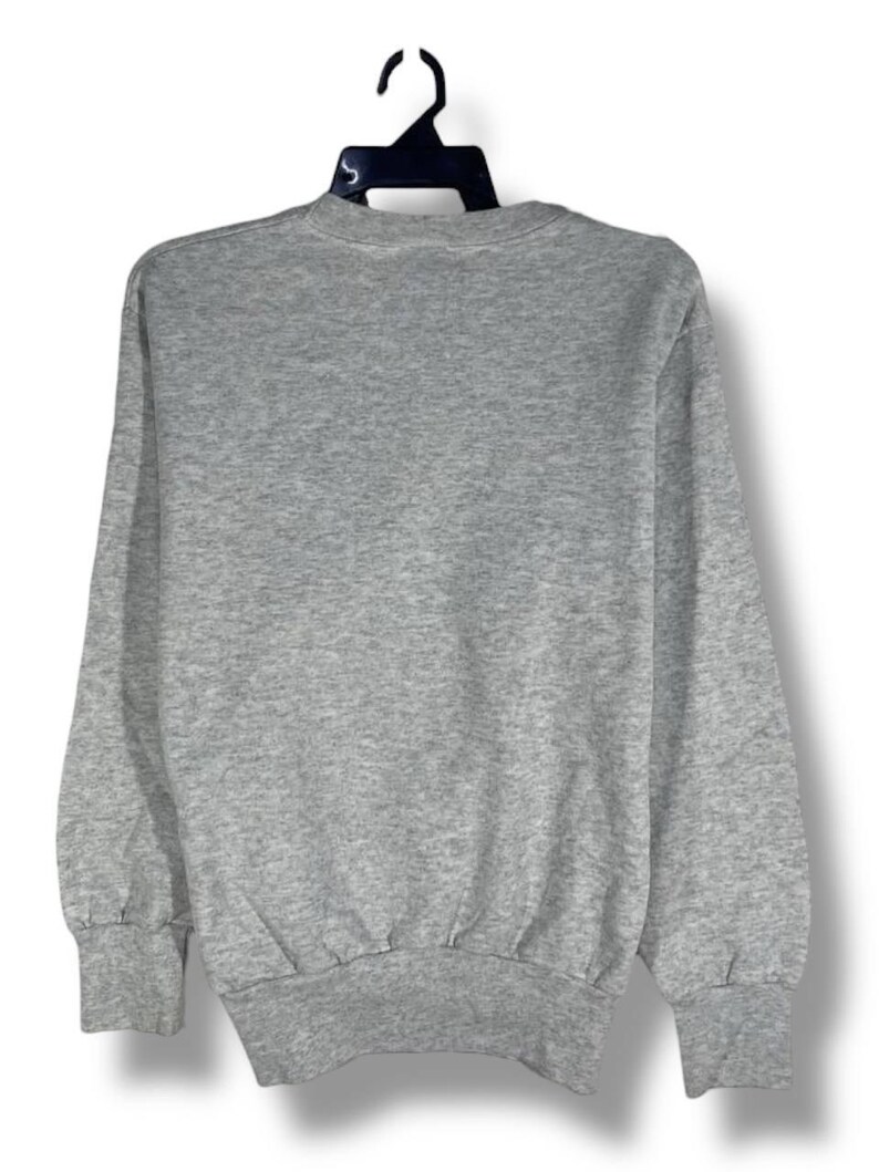 Puede incluir: Una sudadera gris jaspeado con cuello redondo y mangas largas. Los pu&ntilde;os y el dobladillo son acanalados. La sudadera est&aacute; colgada de una percha negra sobre un fondo blanco. Se ve la parte trasera de la sudadera.