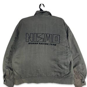 May include: A grey jacket featuring the text "NISMO NISSAN RACING TEAM" in black on the back. The jacket has a collar, long sleeves, and buttoned cuffs. It is suspended from a black hanger.