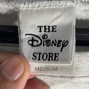 Puede incluir: Primer plano de una etiqueta blanca de The Disney Store. La etiqueta presenta el logotipo de Disney y la palabra "STORE" en texto negro. La palabra "MEDIUM" est&aacute; impresa debajo.
