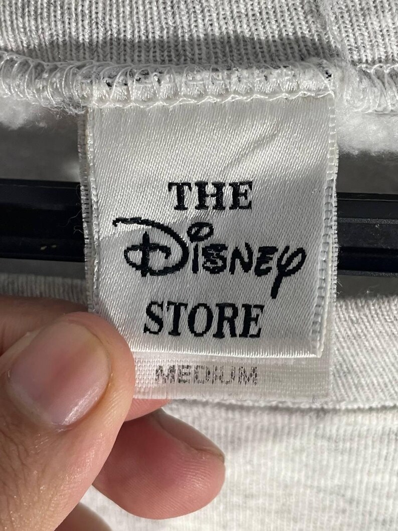 Puede incluir: Primer plano de una etiqueta blanca de The Disney Store. La etiqueta presenta el logotipo de Disney y la palabra "STORE" en texto negro. La palabra "MEDIUM" est&aacute; impresa debajo.