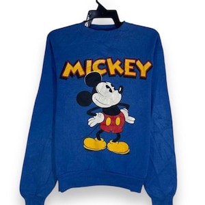 以下が含まれることがあります： 青いスウェットシャツ。ミッキーマウスのグラフィックと「MICKEY」の文字が黄色と赤で描かれています。漫画のキャラクターは、赤いショートパンツ、黄色の靴、白い手袋を身に着けて、象徴的なポーズで描かれています。スウェットシャツは長袖で、クルーネックです。