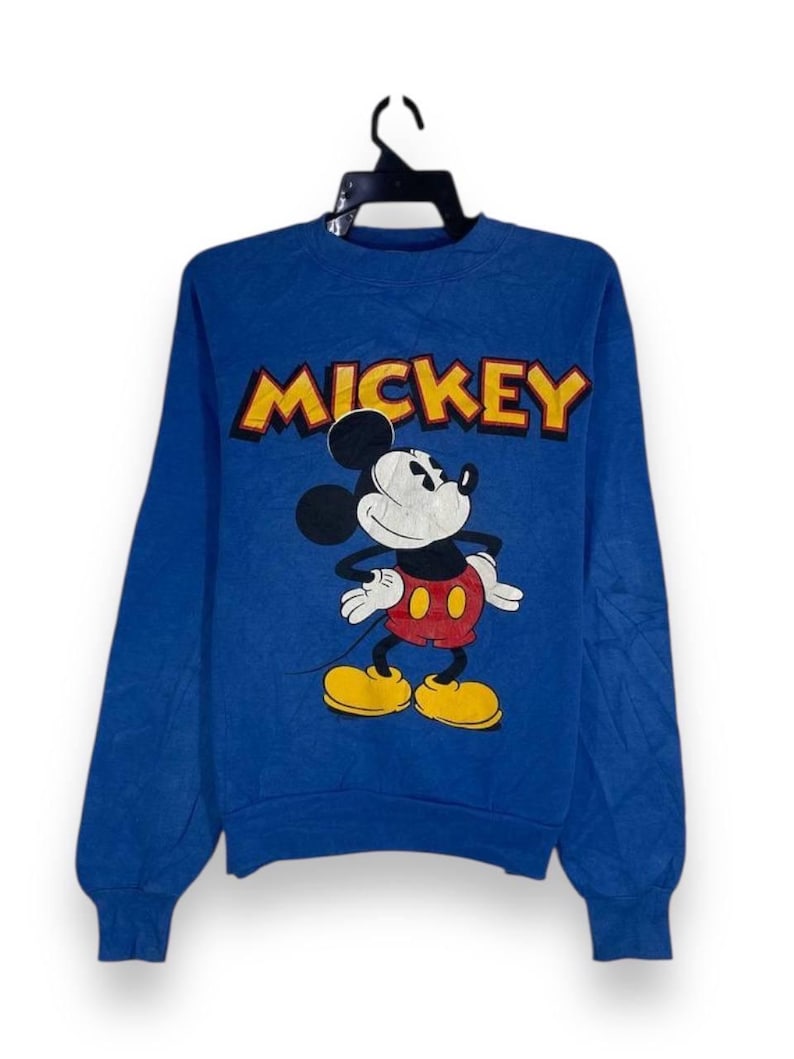 以下が含まれることがあります： 青いスウェットシャツ。ミッキーマウスのグラフィックと「MICKEY」の文字が黄色と赤で描かれています。漫画のキャラクターは、赤いショートパンツ、黄色の靴、白い手袋を身に着けて、象徴的なポーズで描かれています。スウェットシャツは長袖で、クルーネックです。