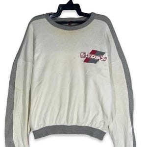 May include: A white long-sleeve sweatshirt with grey trim on the collar, cuffs, and sleeves. The chest features a graphic logo with the text TOM'S in red and black lettering set against a diagonal red and grey block design on a white background.
