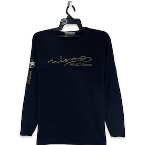 May include: Black long-sleeve shirt with the word "nismo" in gold script across the chest, with "PROJECT DESIGN" below. The left sleeve has a white and gold logo that says "nismo" and "NISSAN RACING".