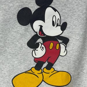 Puede incluir: Una sudadera gris con un gran gr&aacute;fico de Mickey Mouse. Mickey se muestra en su pose cl&aacute;sica, con una cara blanca, orejas negras, brazos negros, pantalones cortos rojos, zapatos amarillos y guantes blancos. El personaje de dibujos animados est&aacute; sonriendo.
