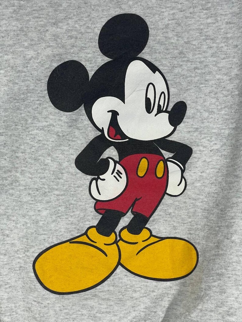 Puede incluir: Una sudadera gris con un gran gr&aacute;fico de Mickey Mouse. Mickey se muestra en su pose cl&aacute;sica, con una cara blanca, orejas negras, brazos negros, pantalones cortos rojos, zapatos amarillos y guantes blancos. El personaje de dibujos animados est&aacute; sonriendo.