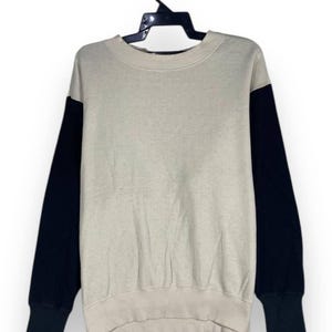 May include: A beige and black colour block sweatshirt hanging on a black hanger. The long sleeves are black, and the body of the sweatshirt is beige. The cuffs and the bottom hem are ribbed.