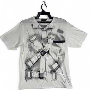May include: A light gray t-shirt featuring a graphic print of a straitjacket. The design includes straps, buckles, and a button-down shirt detail. The shirt is hanging on a black hanger against a white background.