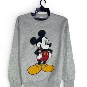 Puede incluir: Sudadera gris con un dibujo de Mickey Mouse. El personaje de dibujos animados est&aacute; representado con su atuendo cl&aacute;sico: orejas negras, guantes blancos, pantalones cortos rojos y zapatos amarillos. La sudadera est&aacute; colgada de una percha negra.