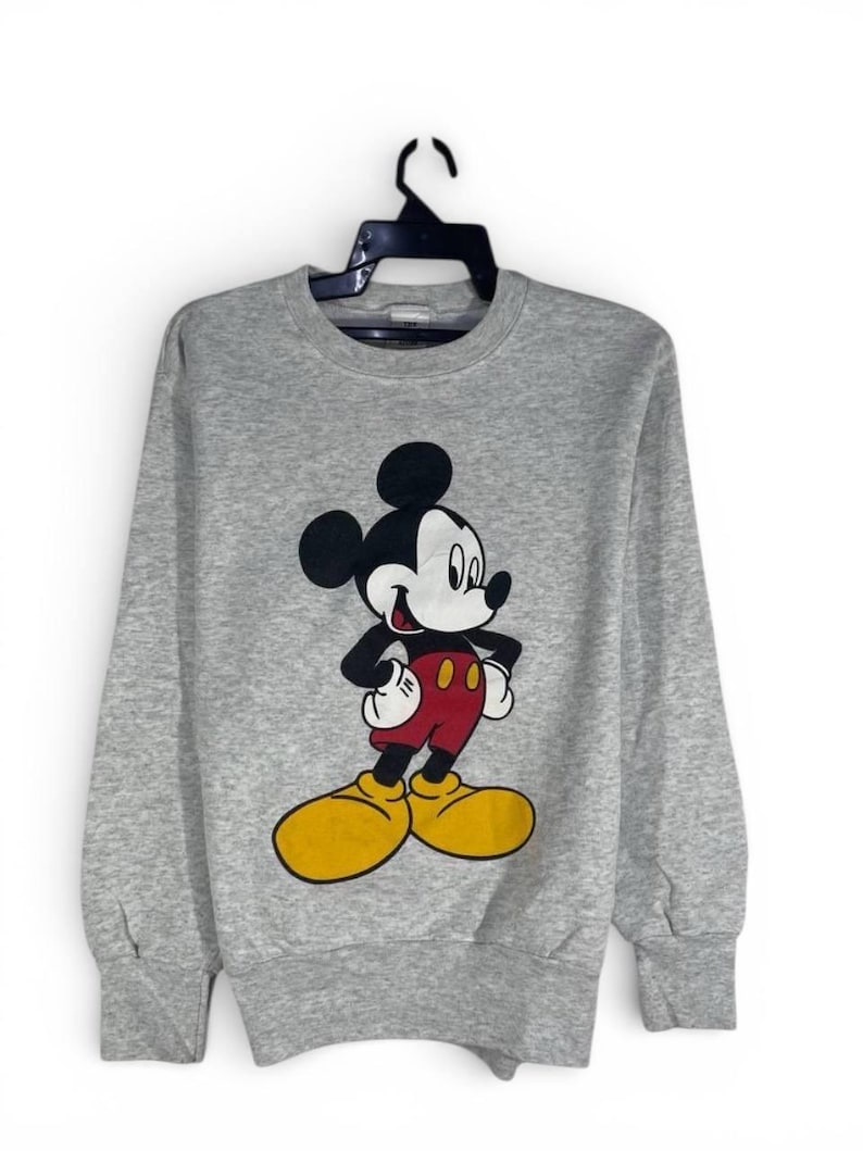 Puede incluir: Sudadera gris con un dibujo de Mickey Mouse. El personaje de dibujos animados est&aacute; representado con su atuendo cl&aacute;sico: orejas negras, guantes blancos, pantalones cortos rojos y zapatos amarillos. La sudadera est&aacute; colgada de una percha negra.