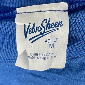 以下が含まれることがあります： 青い衣服のクローズアップで、白いタグが付いています。タグには、ブランド名「Velva Sheen」が様式化されたスクリプトで表示され、「ADULT」とサイズ「M」の文字が下に印刷されています。タグには「OVER FOR CARE MADE IN THE U S A.」とも記載されています。