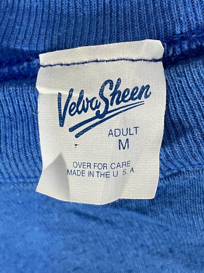 以下が含まれることがあります： 青い衣服のクローズアップで、白いタグが付いています。タグには、ブランド名「Velva Sheen」が様式化されたスクリプトで表示され、「ADULT」とサイズ「M」の文字が下に印刷されています。タグには「OVER FOR CARE MADE IN THE U S A.」とも記載されています。