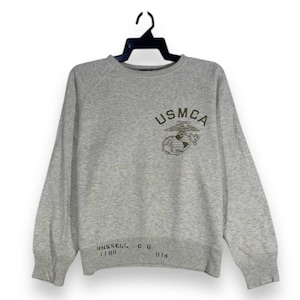 May include: A grey sweatshirt featuring the text "USMCA" and an emblem. The long-sleeved top has a crew neck. Below the emblem are the words "RUSSELL" and "CG" with numbers.