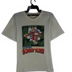 Puede incluir: Camiseta blanquecina con un gráfico de Donkey Kong saliendo de una escena de la jungla. El texto "SUPER DONKEY KONG" está en rojo y amarillo. La camiseta está colgada en una percha negra.