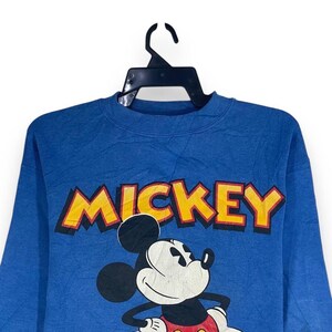以下が含まれることがあります： 黄色い文字で「MICKEY」と書かれた青いスウェットシャツ。ミッキーマウスのクラシックなポーズのグラフィックが特徴で、白い顔、黒い耳、赤いショートパンツが描かれています。黒いハンガーに吊るされています。
