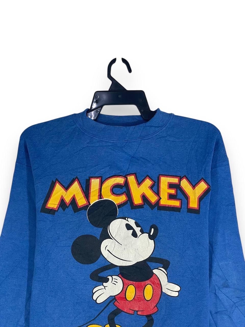 以下が含まれることがあります： 黄色い文字で「MICKEY」と書かれた青いスウェットシャツ。ミッキーマウスのクラシックなポーズのグラフィックが特徴で、白い顔、黒い耳、赤いショートパンツが描かれています。黒いハンガーに吊るされています。