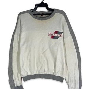 May include: White sweatshirt with grey sleeves and trim. The sweatshirt features the text "TOMS" in red and grey on the front. The garment is hanging on a black hanger.