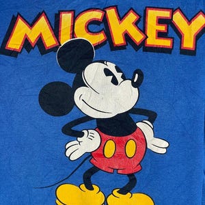 以下が含まれることがあります： ミッキーマウスのグラフィックが描かれた青いTシャツ。「MICKEY」の文字は、キャラクターの上に黒いアウトラインで黄色で印刷されています。ミッキーマウスは、赤いショートパンツ、黄色の靴、白い手袋を身に着けています。