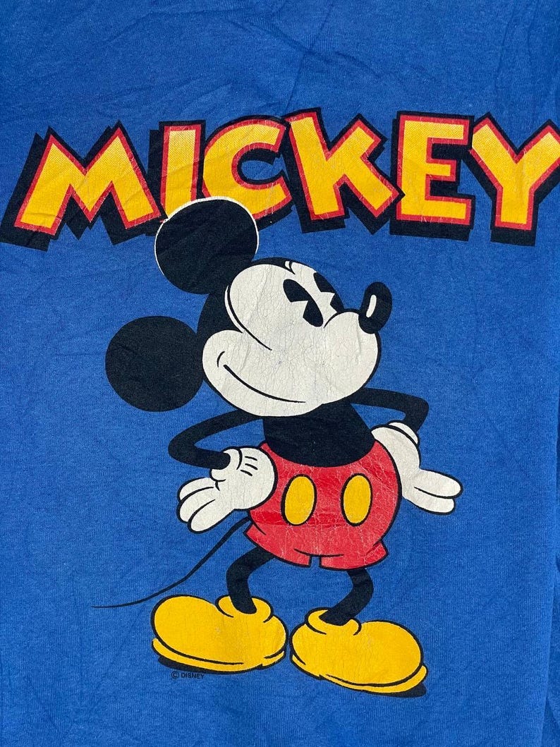 以下が含まれることがあります： ミッキーマウスのグラフィックが描かれた青いTシャツ。「MICKEY」の文字は、キャラクターの上に黒いアウトラインで黄色で印刷されています。ミッキーマウスは、赤いショートパンツ、黄色の靴、白い手袋を身に着けています。