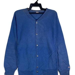 May include: A blue button-up sweatshirt with a V-neck and long sleeves. The sweatshirt has a ribbed hem and cuffs, two front pockets, and a small logo on the left sleeve. It is hanging on a black hanger.