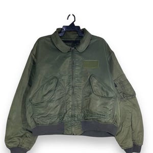 Puede incluir: Chaqueta bomber verde oliva con cremallera, dos bolsillos delanteros grandes y un pequeño parche en el pecho. La chaqueta tiene un dobladillo y puños de color gris oscuro acanalados. El cuello es redondo. La chaqueta está colgada de una percha negra.