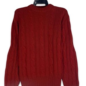 Può includere: Un maglione a maniche lunghe con girocollo di colore rosso scuro. Il capo presenta un motivo a trecce verticali su tutta la parte anteriore, con polsini a coste sui polsi e un orlo a coste sul fondo, il tutto esposto su una gruccia di plastica nera.