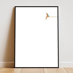 Könnte beinhalten: Gerahmter Kunstdruck mit minimalistischer Illustration. Eine Person in blauem Badeanzug sitzt am Rand einer Holzplattform vor weißem Hintergrund. Das Kunstwerk ist in einem schwarzen Rahmen, geeignet für Wanddekoration.