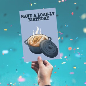 May include: A light blue greeting card featuring a dark grey Dutch oven with a loaf of bread inside and the text HAVE A LOAF-LY BIRTHDAY in bold black letters. The illustration shows steam rising from the bread and the lid resting beside the pot.