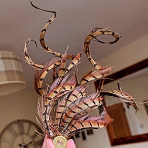 Puede incluir: Arreglo decorativo de plumas marrones y beige con rayas negras y toques de rosa. Las plumas están unidas a una vaina dorada de cartucho de escopeta. El arreglo tiene una estética natural y orgánica.