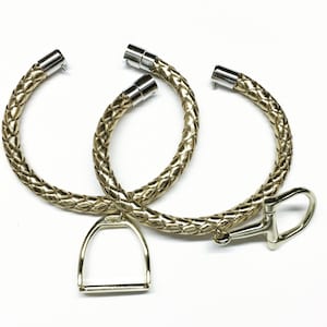 Puede incluir: Dos pulseras de cuero trenzado dorado con cierres plateados y un dije de estribo de metal dorado.