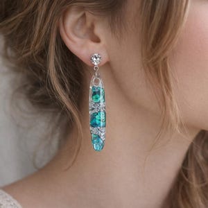 May include: A close-up of a woman wearing a long, dangling earring. The earring features a clear, rectangular resin design with teal and silver accents. The earring is attached to a small, round, clear stud with a crystal.