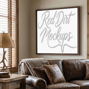 May include: A framed canvas with the text "Red Dirt Mockups" in a cursive font. The frame is dark brown, and the canvas is white. The canvas is hanging above a brown leather sofa with pillows and a blanket.