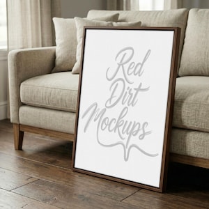 Puede incluir: Una maqueta de lienzo enmarcada con el texto "Red Dirt Mockups" en cursiva. El marco es marrón oscuro y el lienzo es blanco. La maqueta está apoyada contra un sofá beige sobre un suelo de madera.