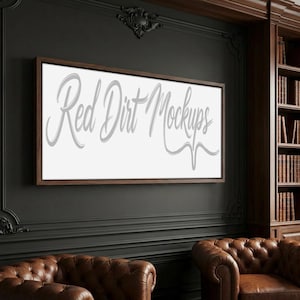 May include: A framed white canvas with the text "Red Dirt Mockups" in a cursive font. The frame is a dark brown color. The canvas is mounted on a dark gray wall, with two brown leather armchairs in the foreground.