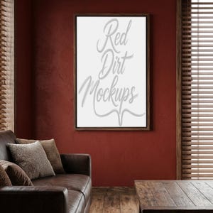 Puede incluir: Un lienzo enmarcado con el texto "Red Dirt Mockups" en fuente cursiva. El marco es marrón oscuro, sobre un fondo rojo intenso. Un sofá de cuero marrón y una mesa de madera están en primer plano.