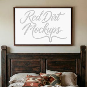 Op de afbeelding: Een ingelijst canvas met de tekst "Red Dirt Mockups" in een sierlijk lettertype hangt boven een bed met decoratieve kussens. Het bedframe is van donker hout en de kussens hebben patronen in rood, bruin en groen.