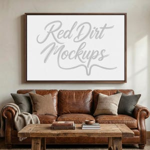 May include: A framed canvas with the text "Red Dirt Mockups" in a cursive font hangs above a brown leather sofa. The sofa has several throw pillows and a wooden coffee table in front of it.