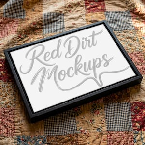 Puede incluir: Un lienzo enmarcado en negro con el texto "Red Dirt Mockups" en fuente cursiva. El lienzo descansa sobre un edredón de retazos con varios patrones y colores.